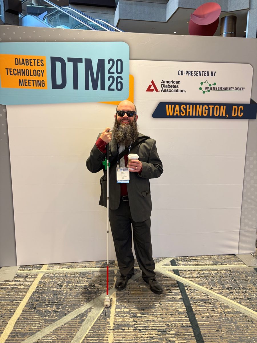 Ismael Collazo standing in front of the DTM 2026 Diabetes Technology Meeting backdrop in Washington, DC, wearing a suit, holding a coffee, white cane in hand.